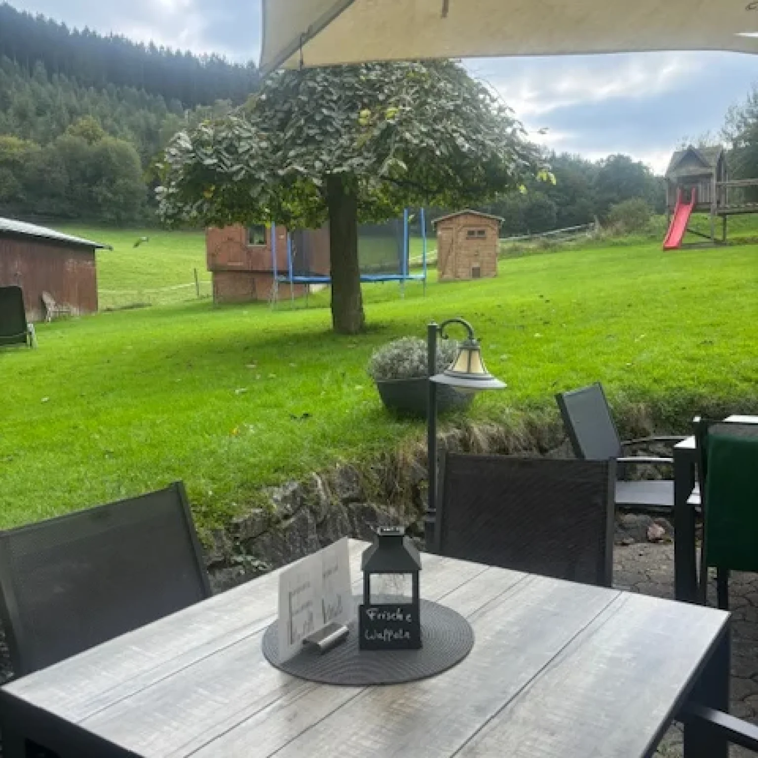 Holztisch mit einer Laterne und einer Menükarte auf einer Terrasse in einer ländlichen Umgebung.