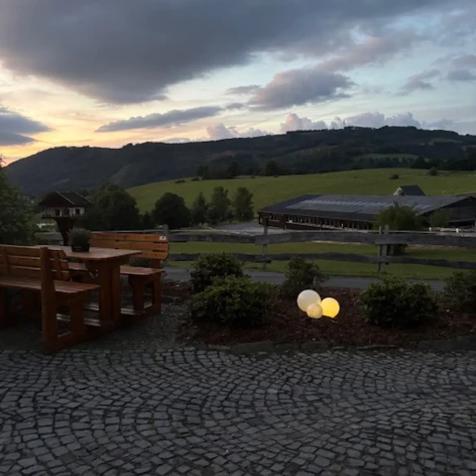 Sitzgelegenheit aus Holz mit Beleuchtung im Vordergrund und Landschaft im Hintergrund bei Sonnenuntergang.
